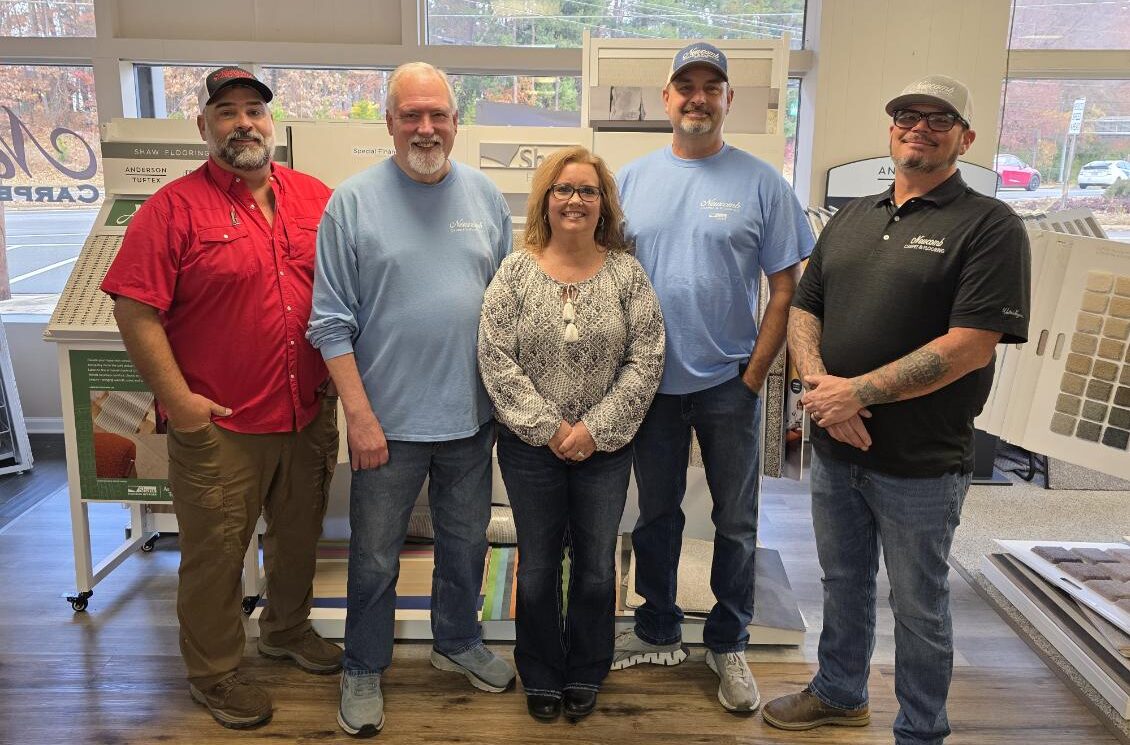 A group of five flooring specialists posing together inside a bright, well-lit showroom featuring carpet and hardwood flooring samples.